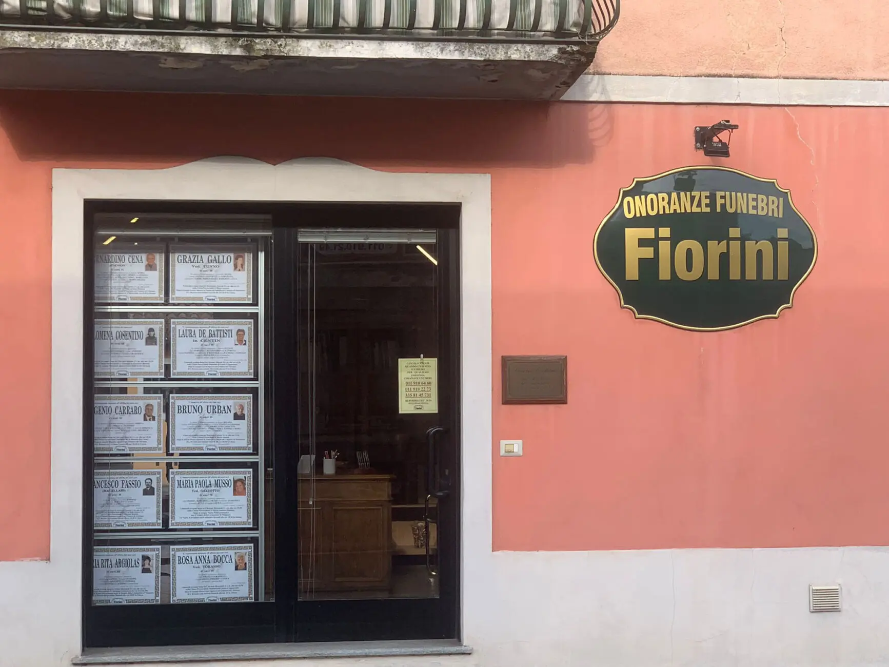 Facciata rosa con insegna verde e oro Onoranze Funebri Fiorini, necrologi in vetrina, porta a vetri sull'ufficio