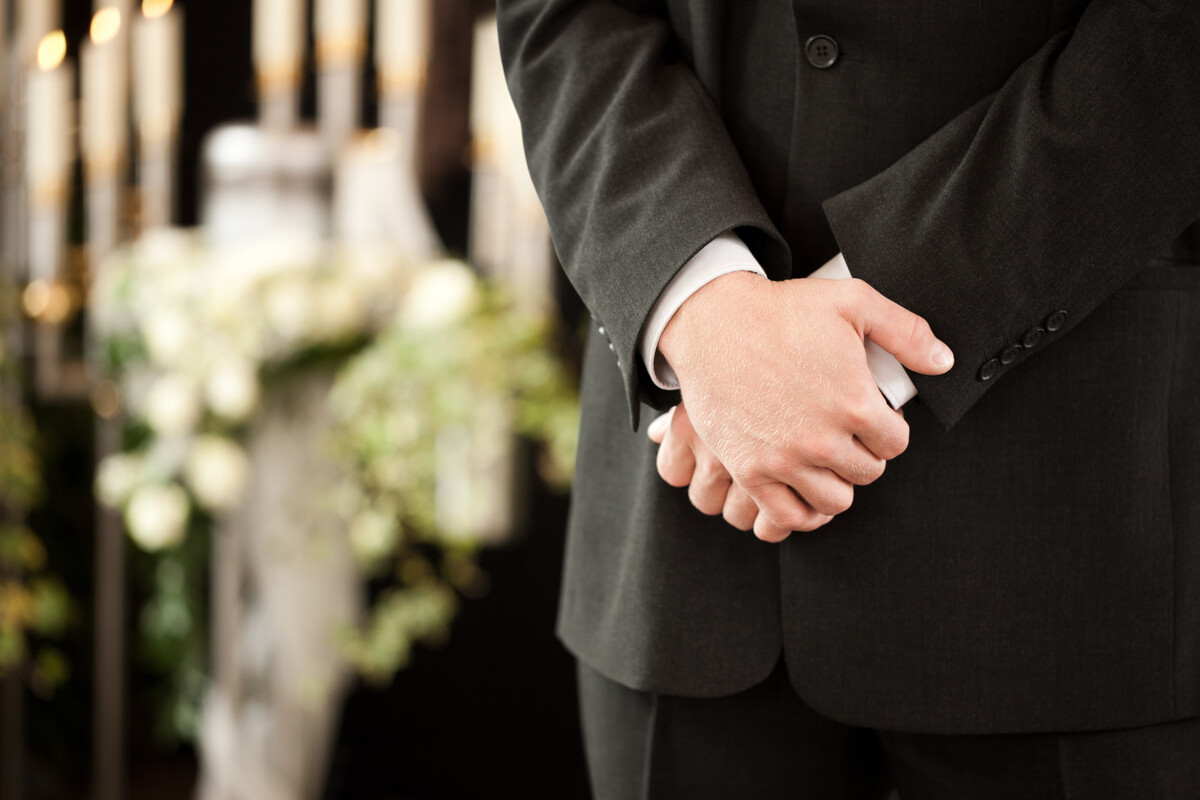 Uomo in abito scuro con le mani giunte davanti a sé, sullo sfondo sfocato fiori bianchi e candele accese