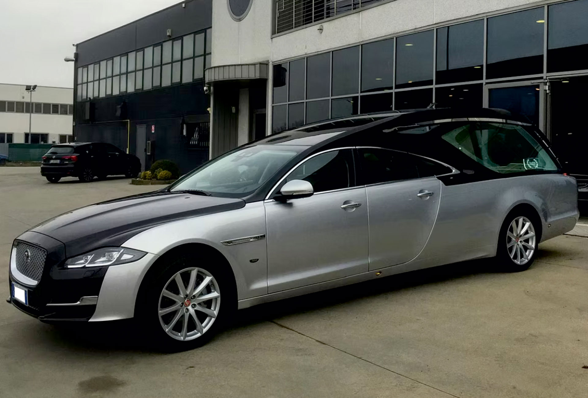 Carro funebre Jaguar argento con cofano e tetto neri parcheggiato davanti a un edificio moderno a vetrate